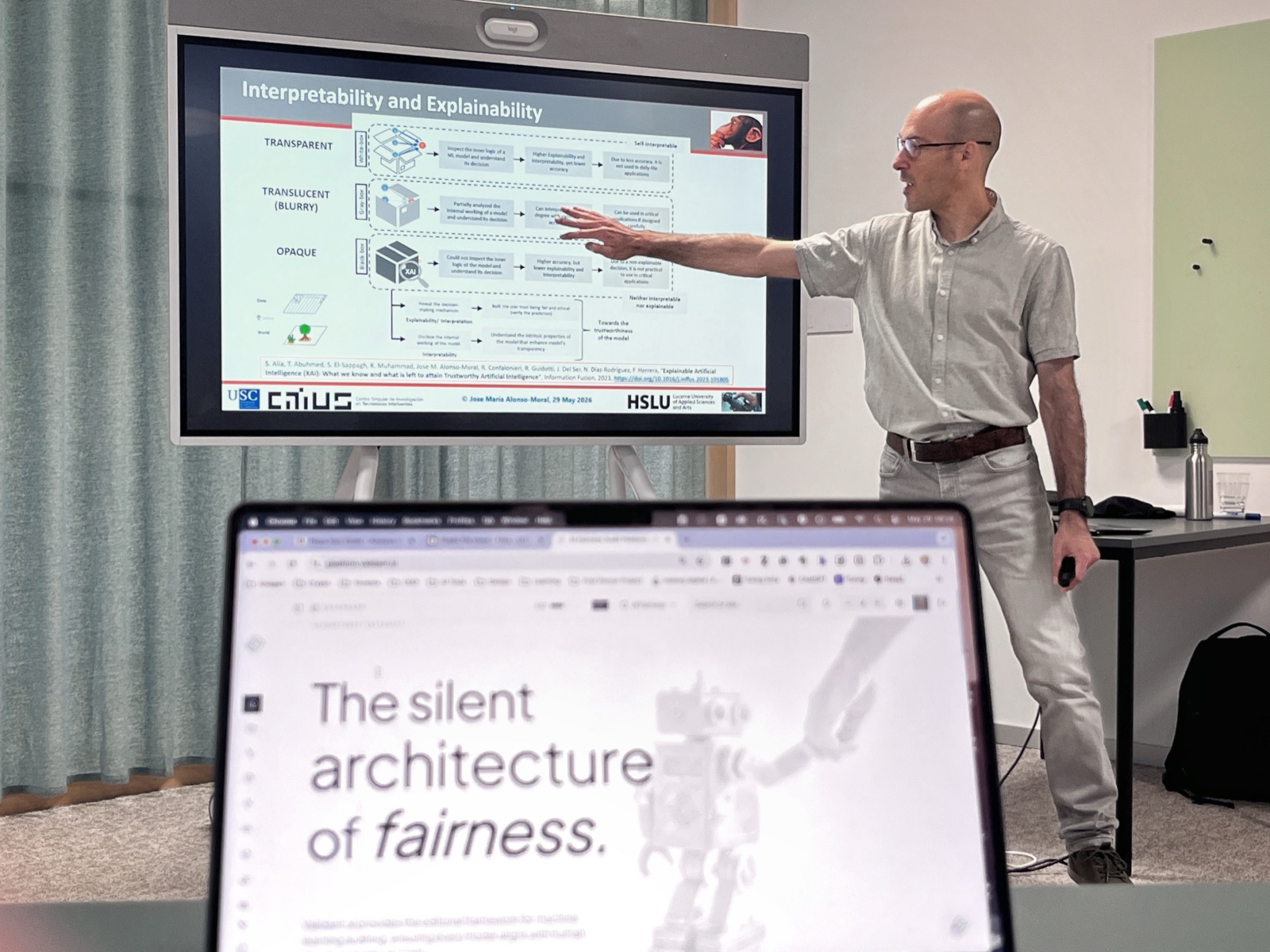 Prof. José María Alonso Moral presenting a slide headed “Interpretability and Explainability”, mapping transparent, translucent and opaque models, while a laptop in the foreground shows the validant.ai page reading “The silent architecture of fairness.”