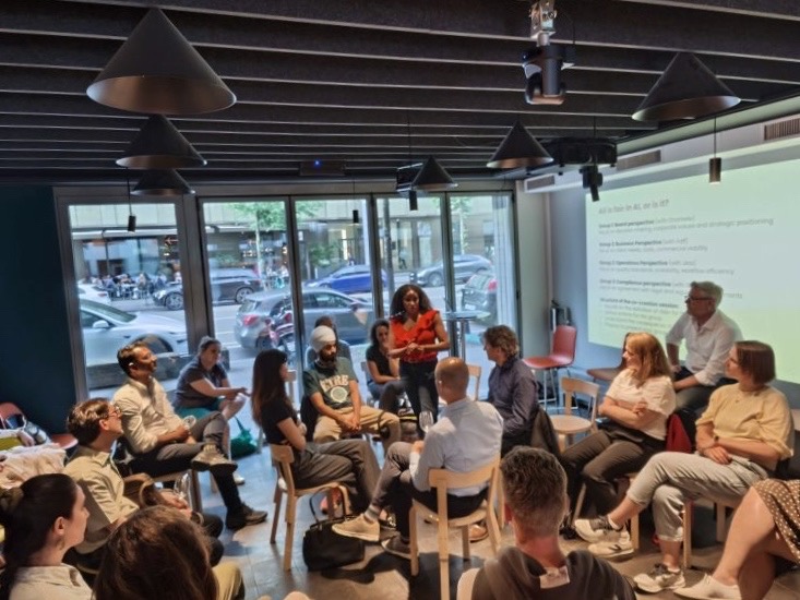 Participants seated in a circle in open discussion, with a projected slide reading “All is fair in AI, or is it?” behind them.