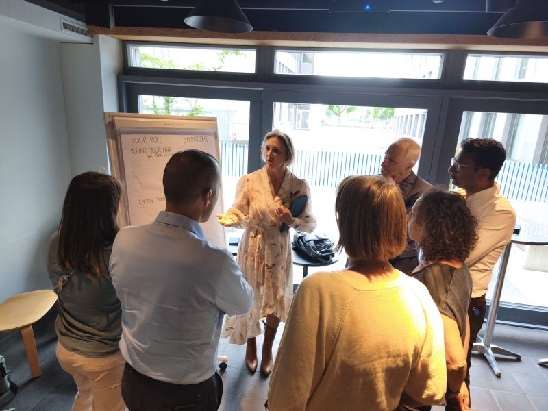 A facilitator presenting at a flipchart headed “Your role: Operations. Define your fair” to a circle of standing participants.